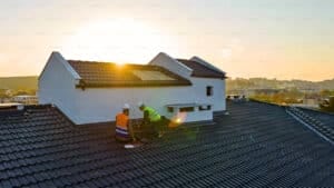Two construction workers wearing safety vests and helmets work on the roof of a house at sunset, performing essential Autumn Roof Maintenance as sunlight shines between two gables, with a view of distant buildings and hills in the background.